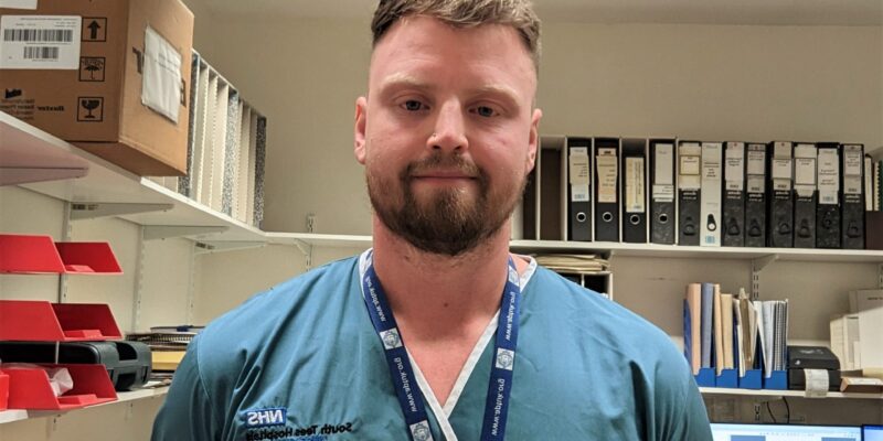 Photo of Curtis Booth stood in full uniform (green scrubs) with his hands behind his back.